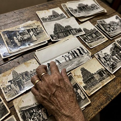 An elderly woman’s hand, with gold rings, resting on old black-and-white photos of temples spread across a wooden table
