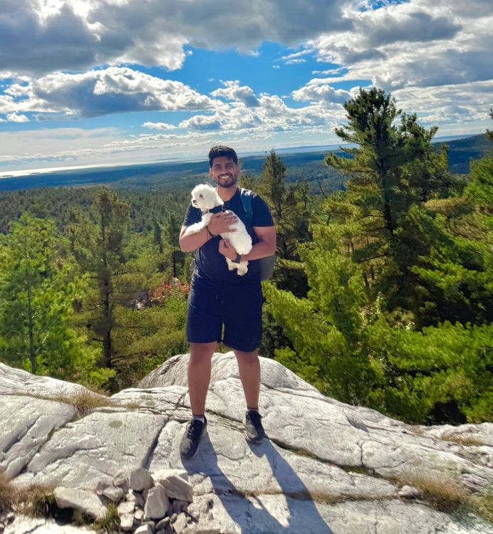 A smiling hiker holds a small white fluffy dog on a granite ridge above a forested valley