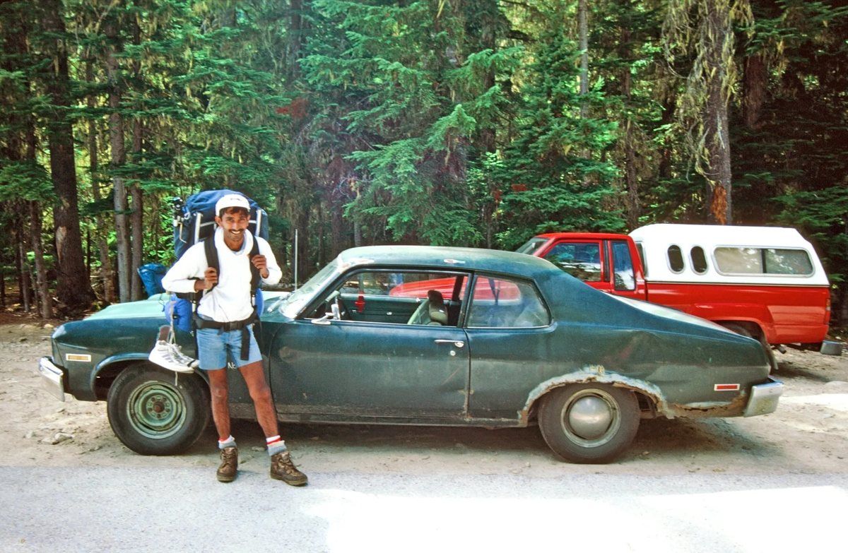 A smiling young man with a big backpack stands beside a rusty green two-door car at a forest trailhead