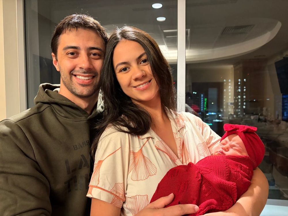 A smiling couple at night by a window, the mother holding their newborn daughter swaddled in red with a red bow