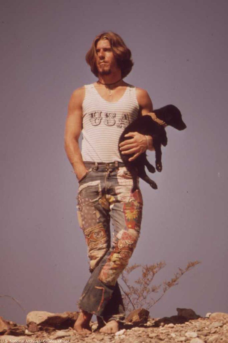 A young man with long hair in a USA tank top and patchwork jeans holds a black puppy in the desert