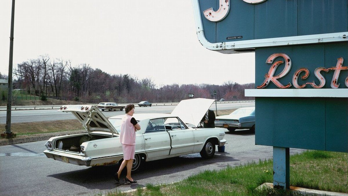 A woman in a pink suit stands by a white Chevrolet with its hood and trunk open beside a roadside restaurant sign
