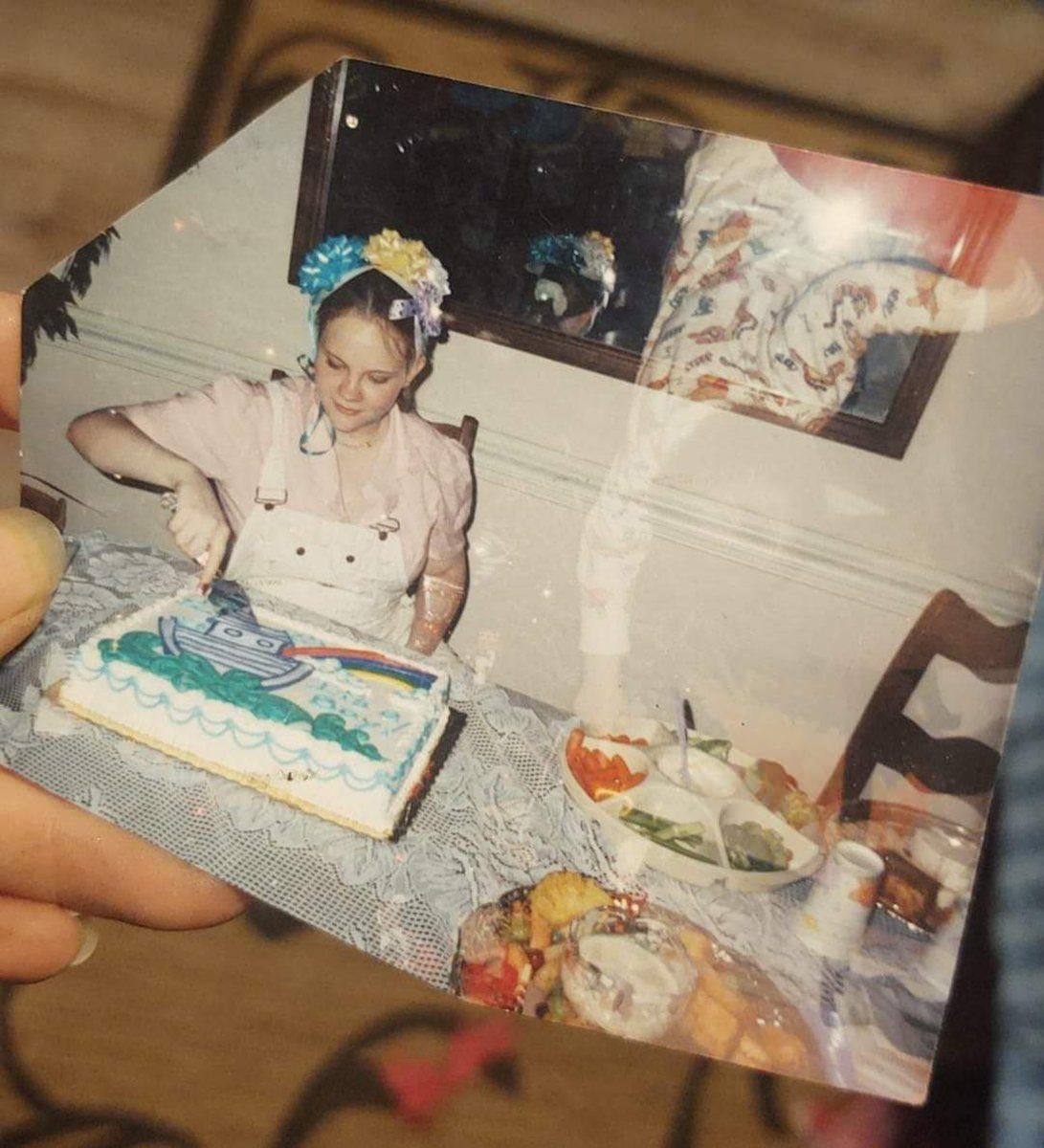 A young woman wearing a hat made of gift bows cuts a Noah’s Ark cake at a table, in a snapshot held in someone’s hand